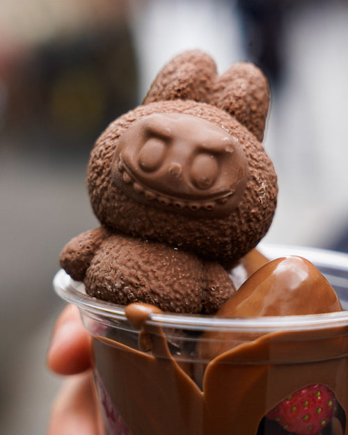 Hand holding a transparent dessert cup filled with melted Dubai chocolate and topped with a solid chocolate Labubu figure