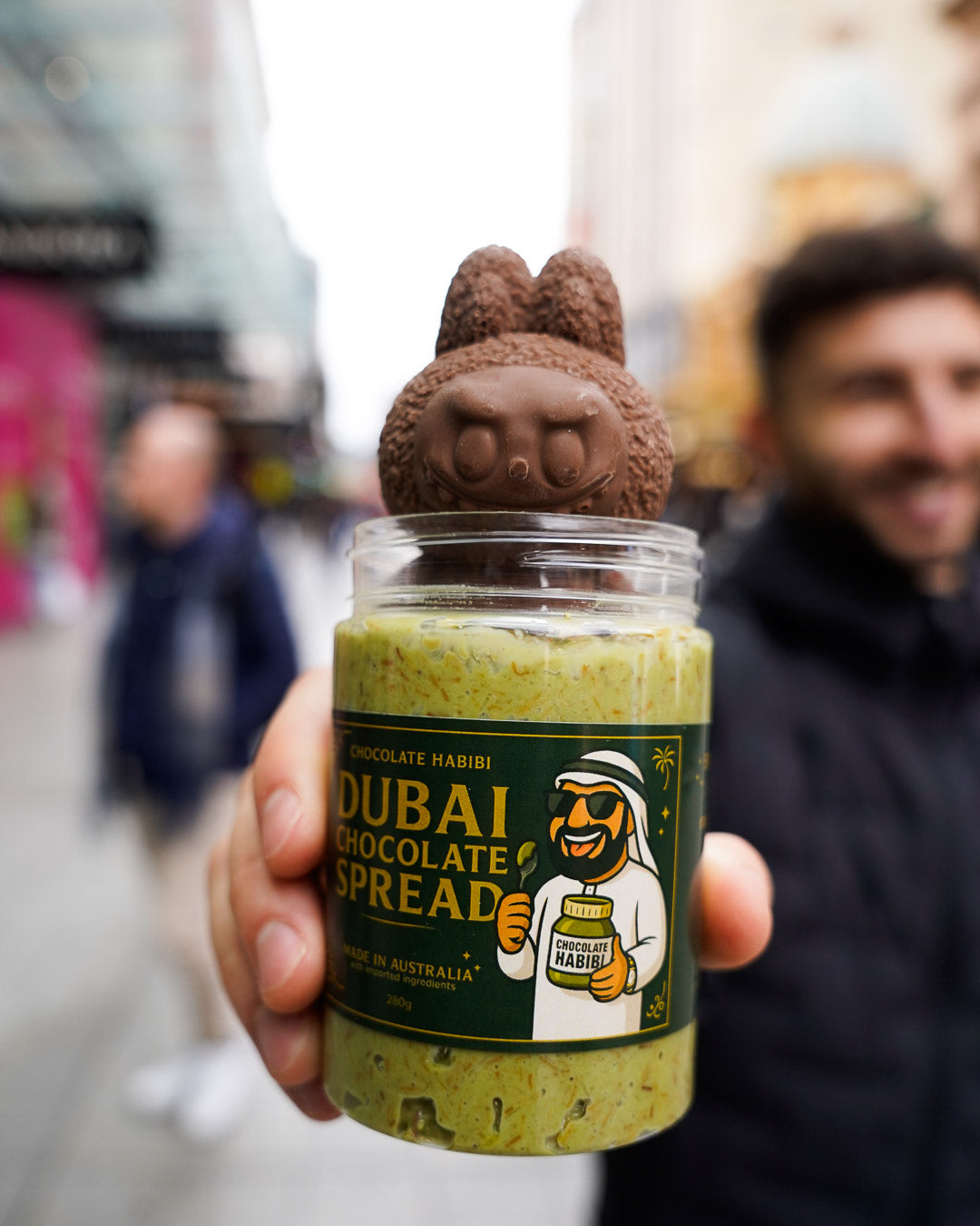 Jar of Dubai Chocolate Spread topped with a chocolate Labubu figure, held by a smiling person in a busy urban street.