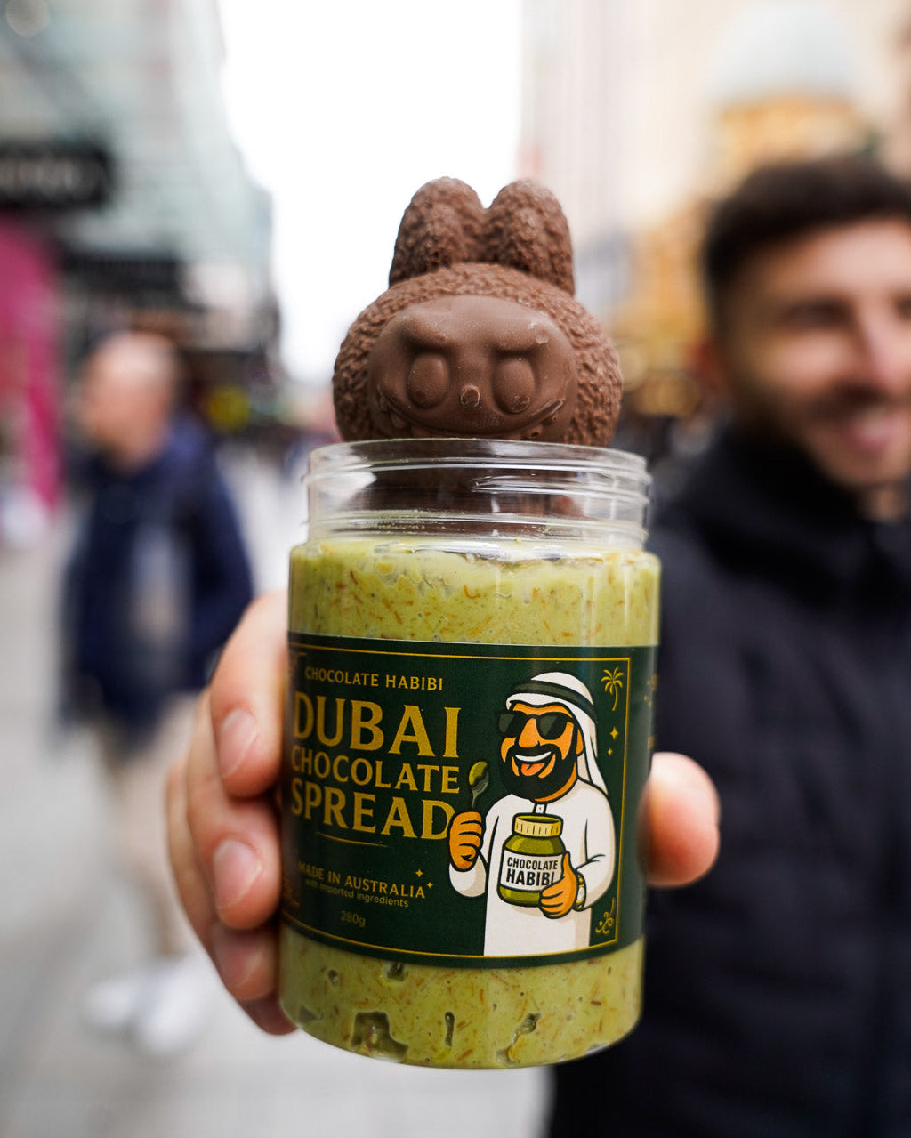 Jar of Dubai Chocolate Spread topped with a chocolate Labubu figure, held by a smiling man on a city street