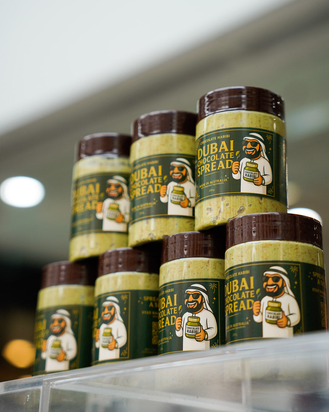 Stacked jars of Dubai Chocolate Spread with pistachio, displayed on a clear shelf with blurred store background