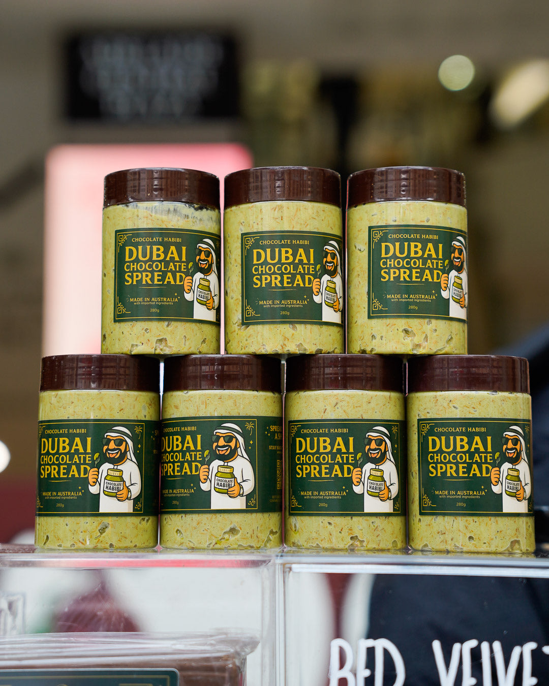 Stacked jars of Dubai Chocolate Spread with pistachio, displayed on a clear shelf with blurred store background