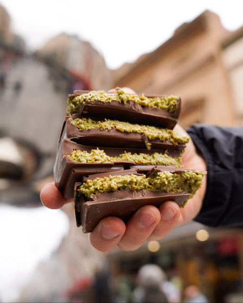 Hand holding a stack of Dubai Pistachio Chocolate bars with vibrant green pistachio filling, against a blurred street background