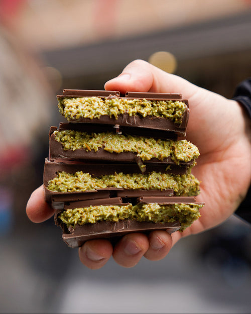 Hand holding a stack of Dubai Pistachio Chocolate bars with vibrant green pistachio filling, against a blurred street background