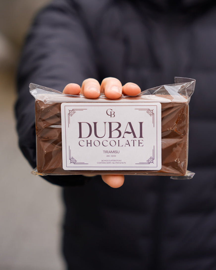 Person holding a sealed Tiramisu Dubai Chocolate bar in clear packaging with a premium label, blurred urban background